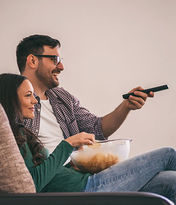 Man with TV remote, and woman eating chips, sat on sofa